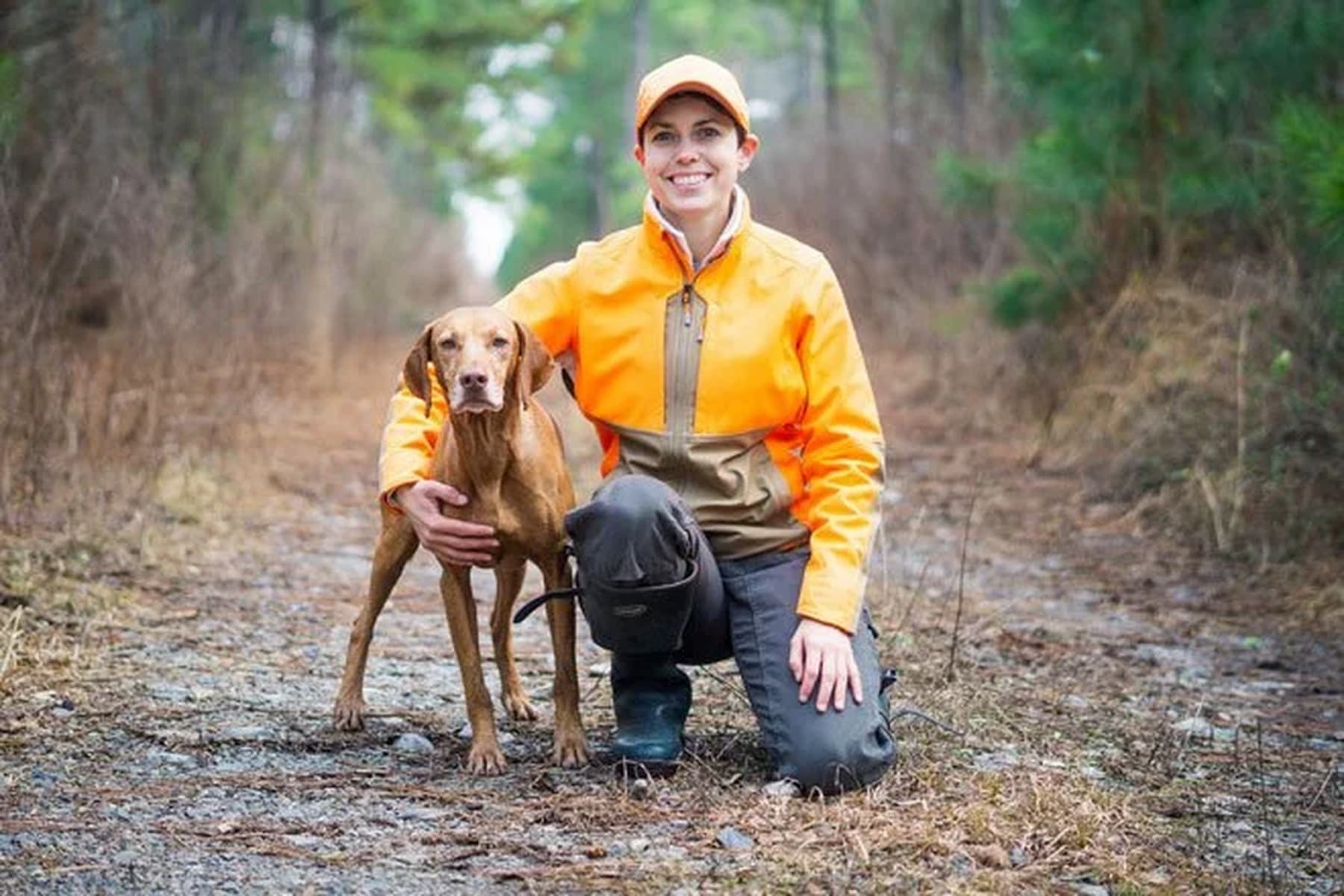 A smiling hunter in an orange jacket kneels beside her pointing dog
